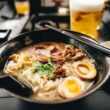 a bowl of ramen with chopsticks and a glass of beer